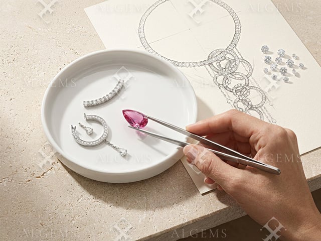 One hand using tweezers to hold a pink pear above a round ivory dish with pavé component studies, beside a graphite necklace drawing and loose clear stones